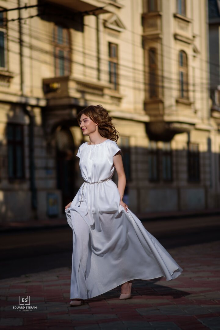 Graceful woman in a flowing white dress walking confidently on a city street, with elegant historic architecture in the background.