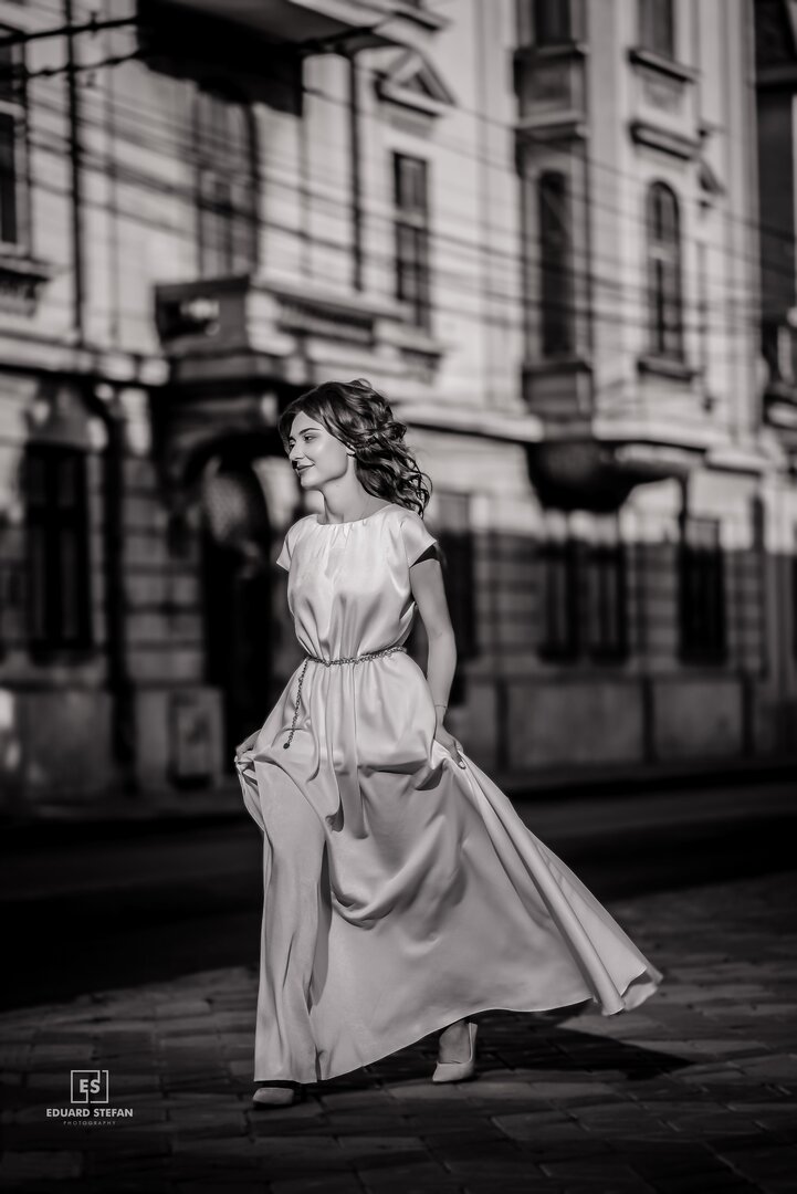 Elegant woman in a flowing white dress captured in black and white, walking on a city street with historic architecture in the background.