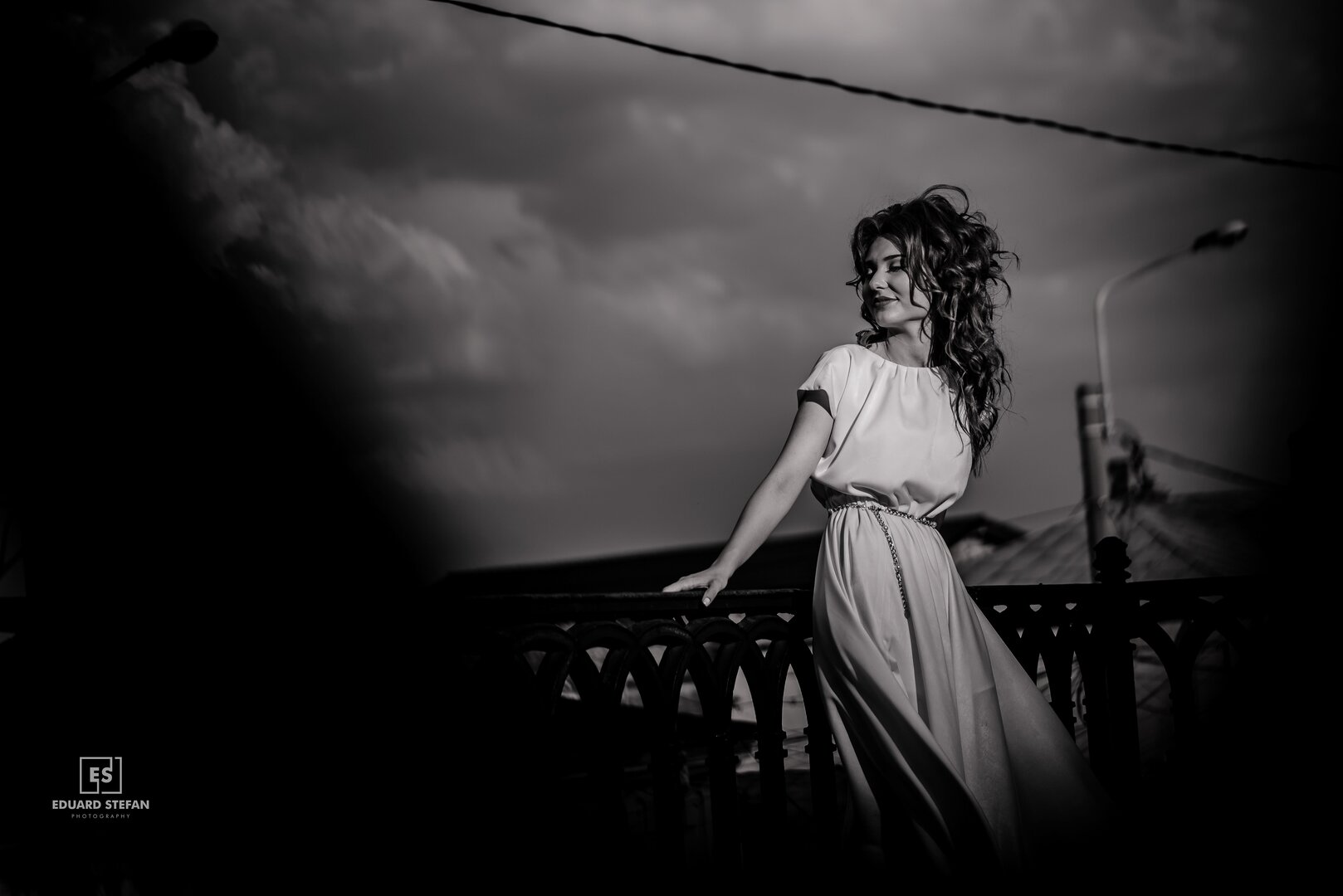 Black and white portrait of a woman in a flowing dress leaning on a railing, with dramatic clouds and urban elements in the background.