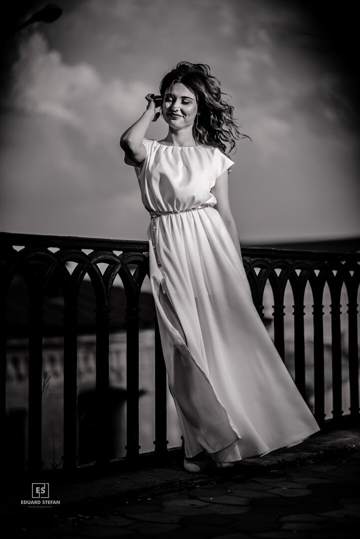 Black and white image of a woman in a flowing dress standing by a decorative railing, with windswept hair and dramatic clouds in the background.