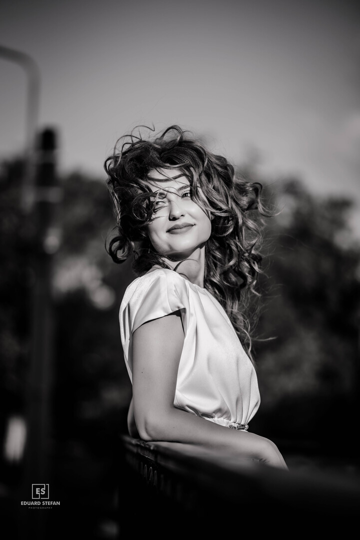 Black and white portrait of a smiling woman with voluminous windswept hair, leaning gracefully on a railing outdoors