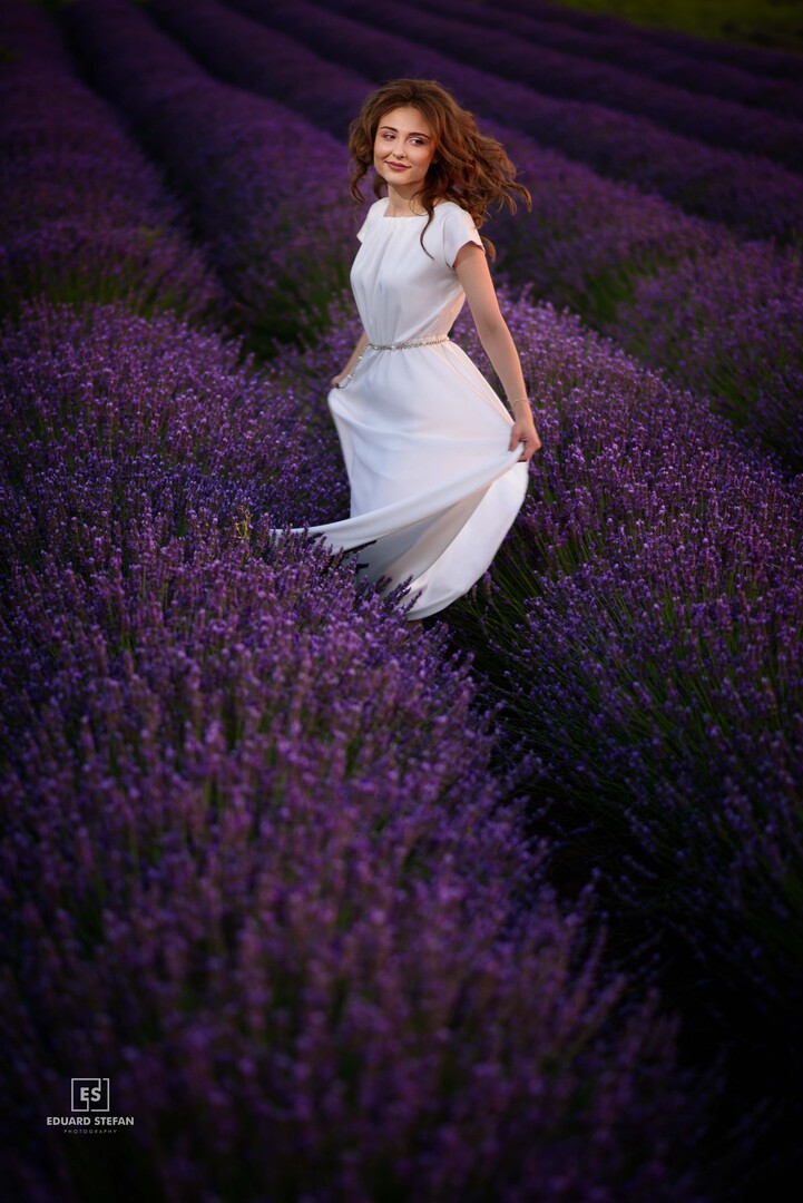 legant woman in a flowing white dress twirls gracefully amidst vibrant purple lavender fields, exuding charm and joy.