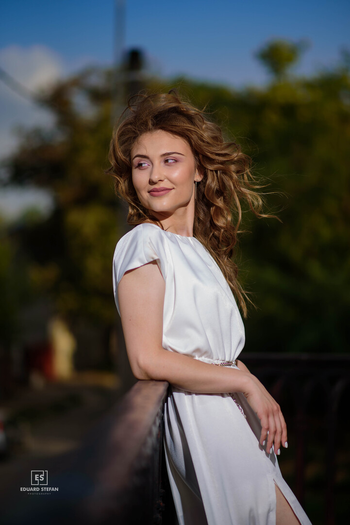 Elegant woman in a flowing white dress leaning on a railing, with her hair gently blown by the breeze in a sunlit outdoor setting.