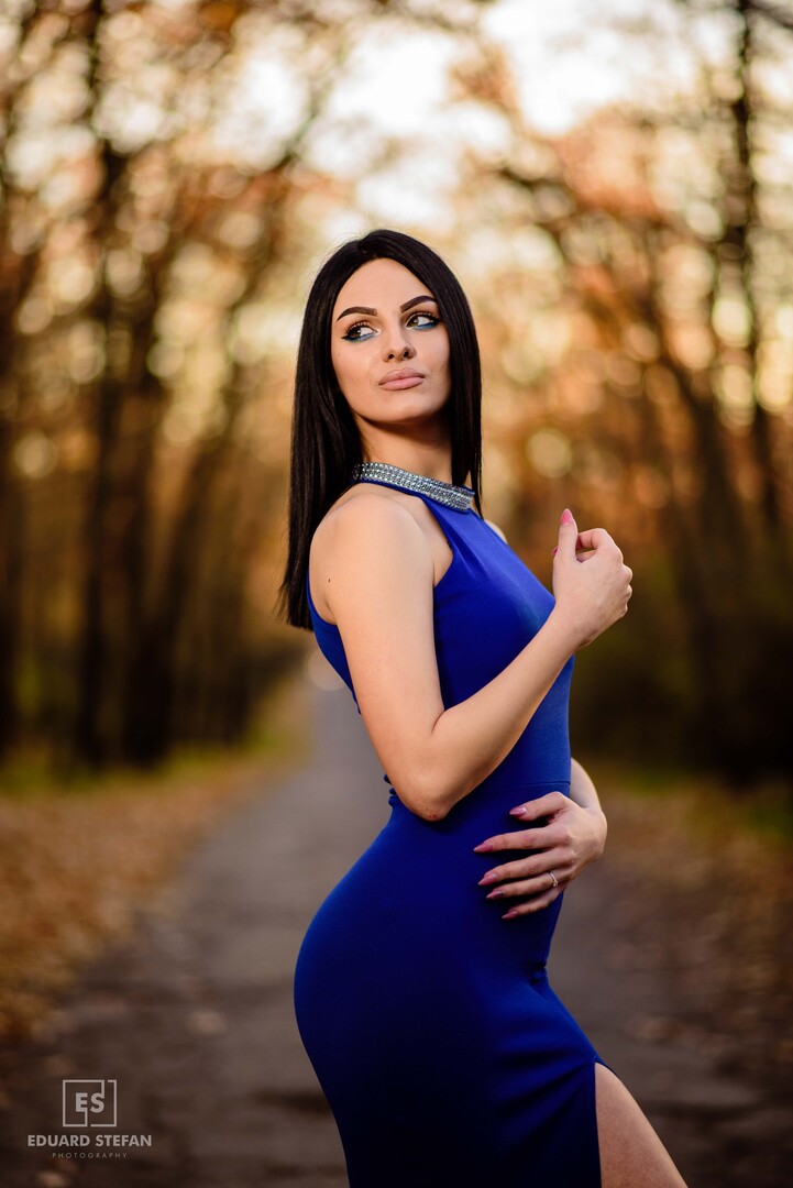 Elegant woman in a striking blue dress standing confidently on a tree-lined pathway, exuding grace and sophistication.