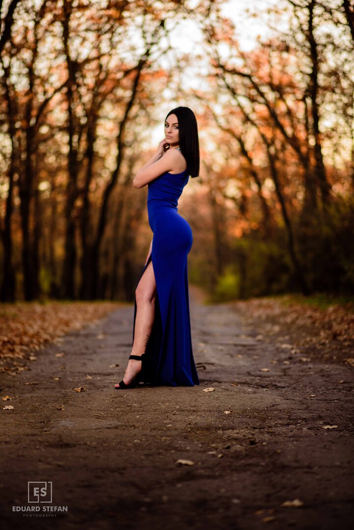 A captivating photo of a woman in a striking blue gown, elegantly poised on an autumn pathway lined with golden trees, showcasing a harmonious blend of nature and fashion.