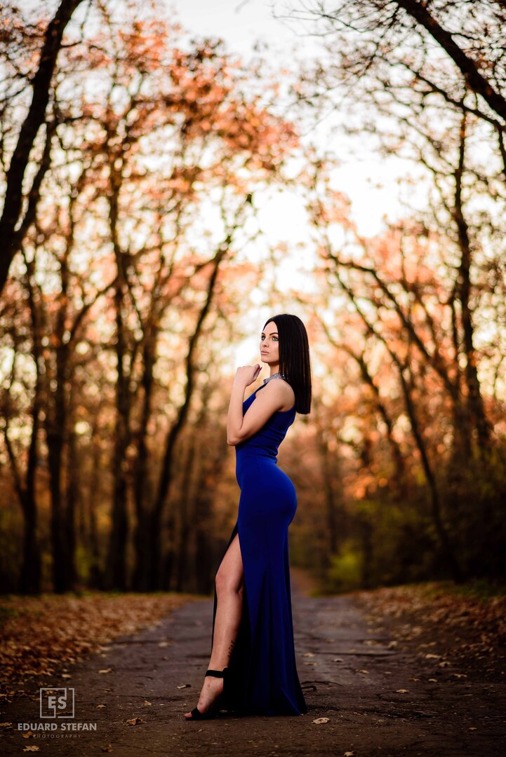 A stunning photograph capturing a woman in an elegant blue gown, posed confidently amidst a picturesque autumn forest, with golden leaves framing the serene pathway.