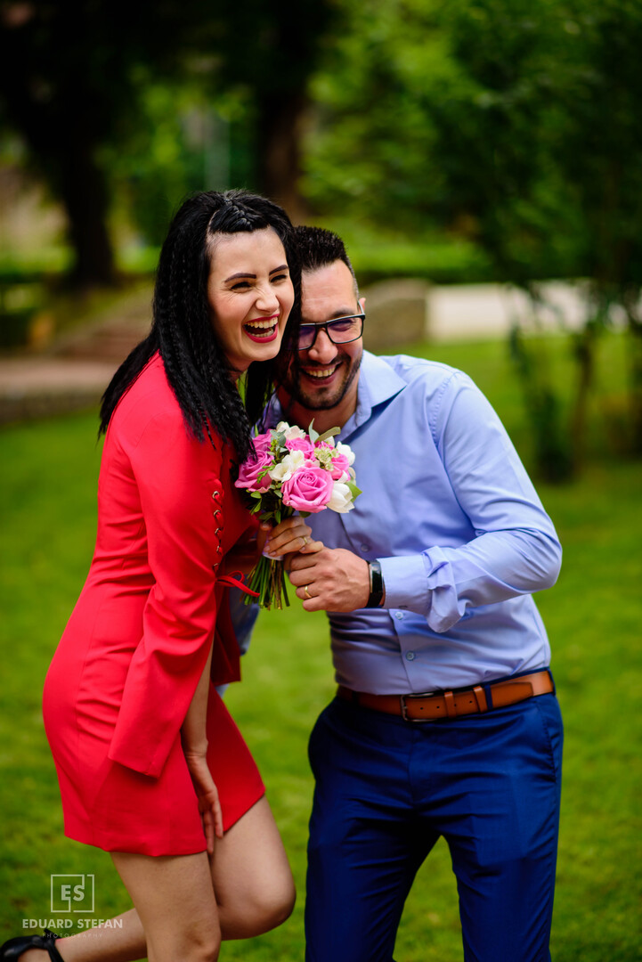 Cheerful couple laughing together outdoors, with the woman holding a bouquet of pink roses.