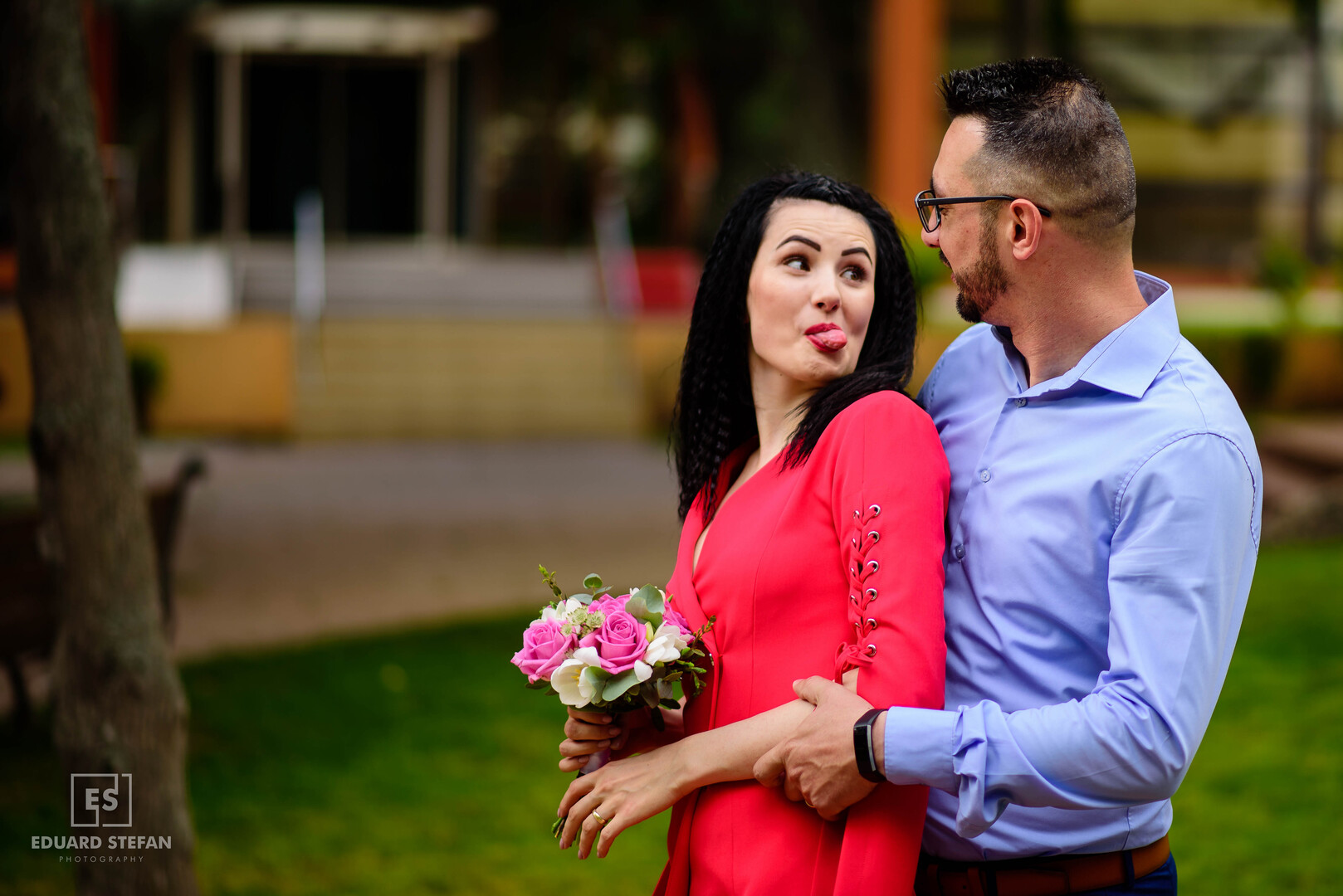 Playful couple outdoors, with the woman in a red dress holding a bouquet of pink roses and sticking her tongue out.