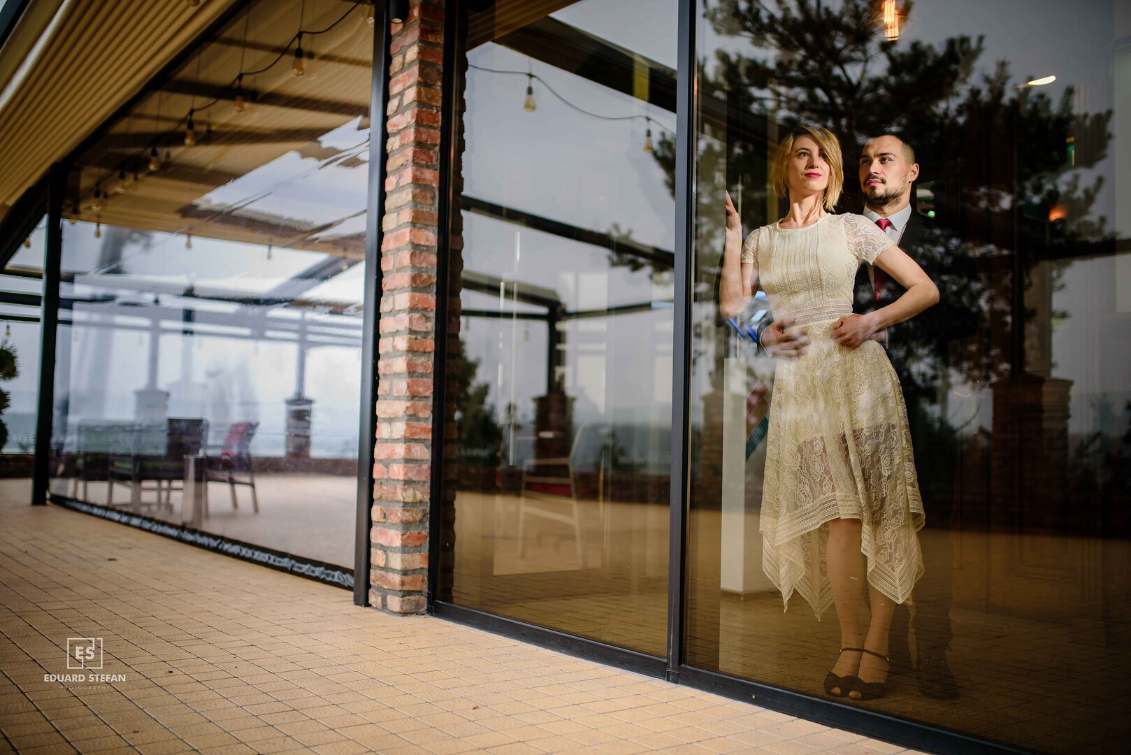 Elegant couple standing behind a glass wall, with their reflection blending into the scenic outdoor patio and trees.