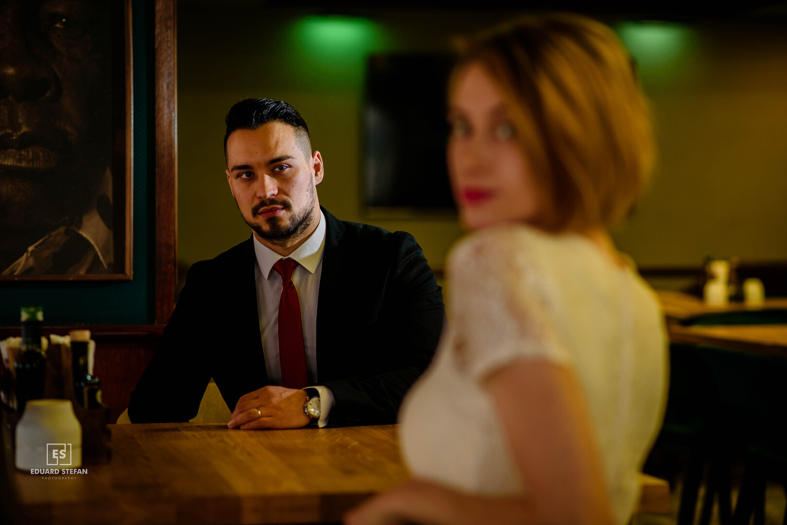 Man in a suit and red tie sitting at a wooden table in a stylish restaurant, with a blurred woman in a white dress in the foreground, captured in a cinematic, moody setting.