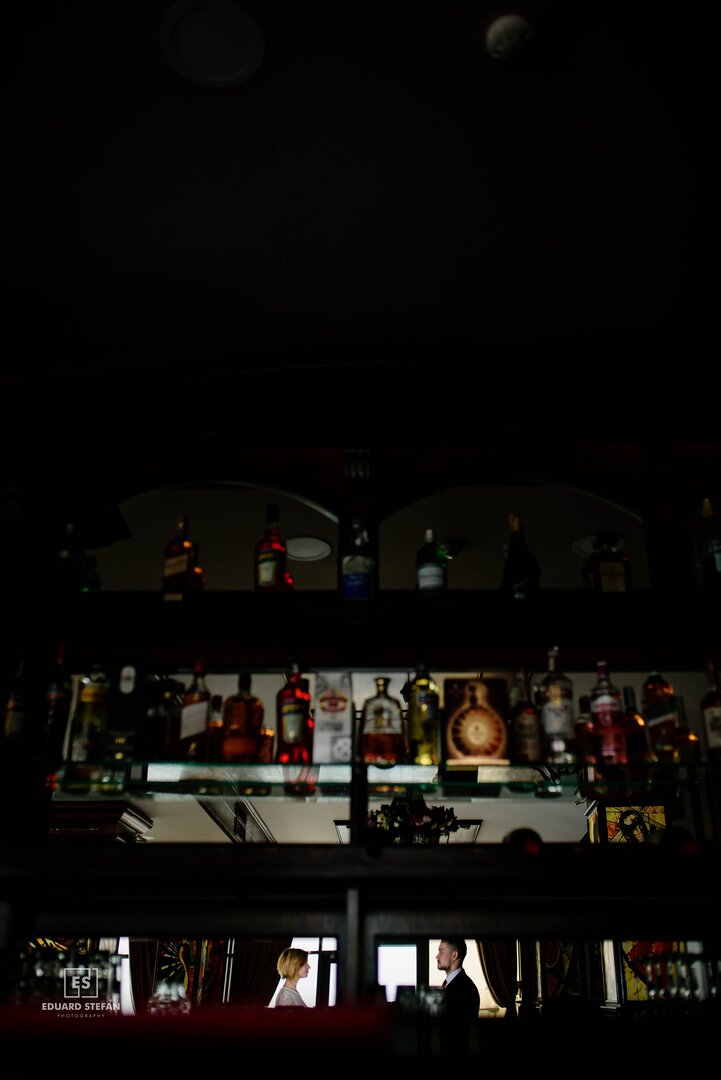 Moody bar interior with a focus on rows of liquor bottles, reflecting a man and woman having a conversation in the background, framed through the bar's glass shelving