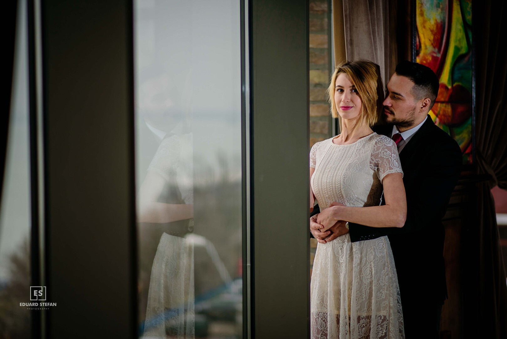 Man in a suit embracing a woman in a white dress near a window, with their reflection visible in the glass.