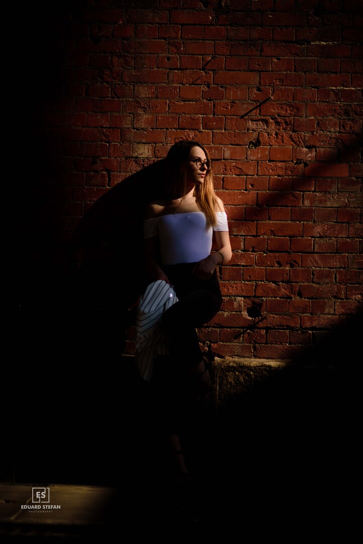 A woman leans against a brick wall, elegantly lit by a striking beam of sunlight amidst deep shadows.