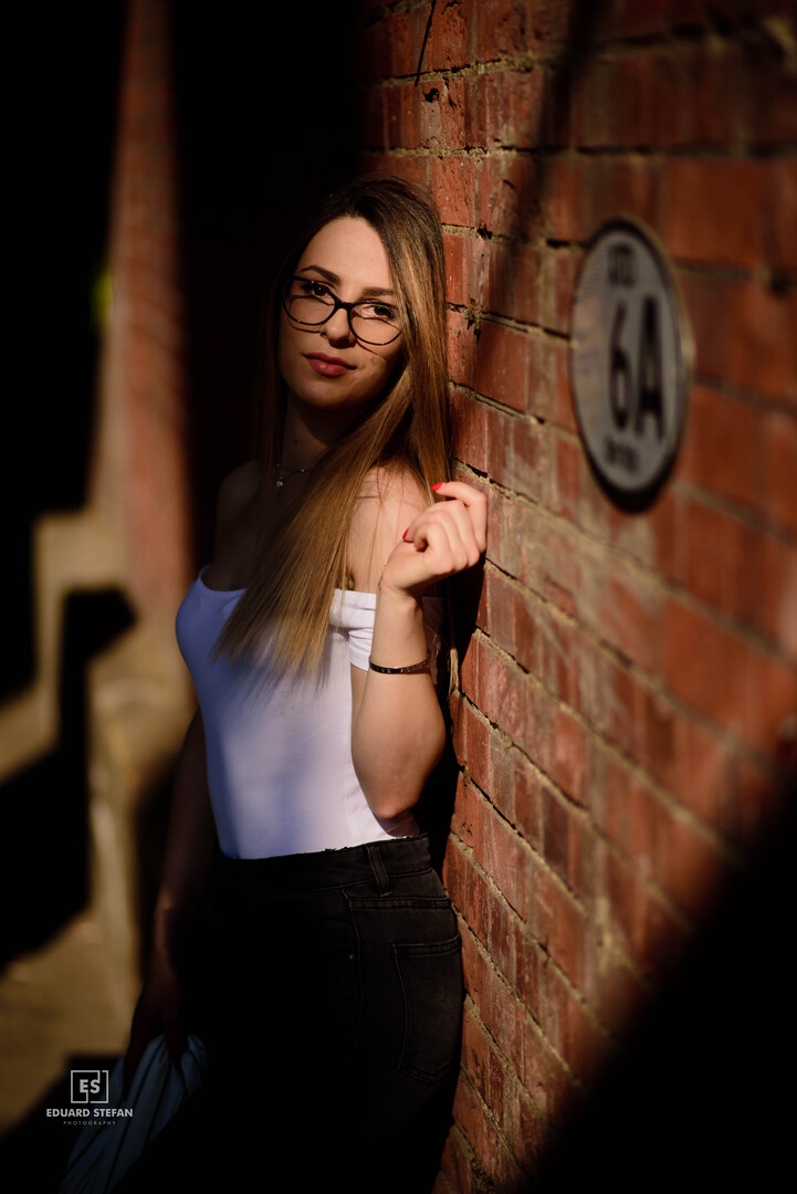 A woman stands against a sunlit brick wall, softly illuminated, her expression calm and confident.