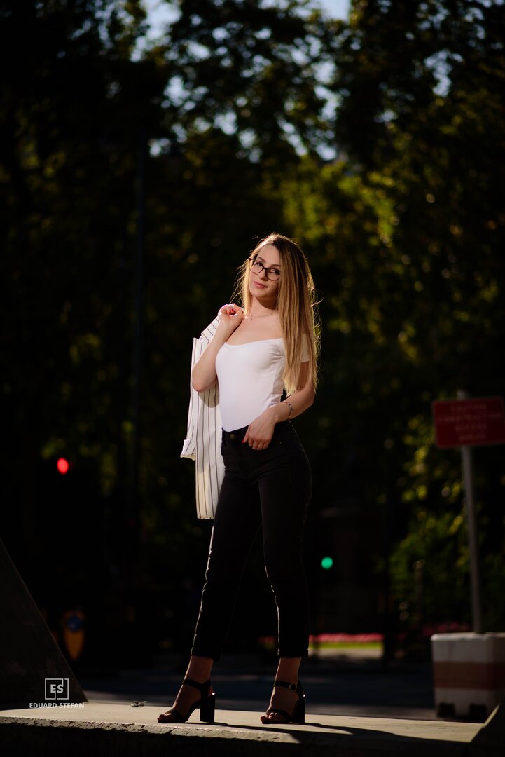 A young woman exudes confidence, standing stylishly on a sunlit urban street with lush greenery in the background.