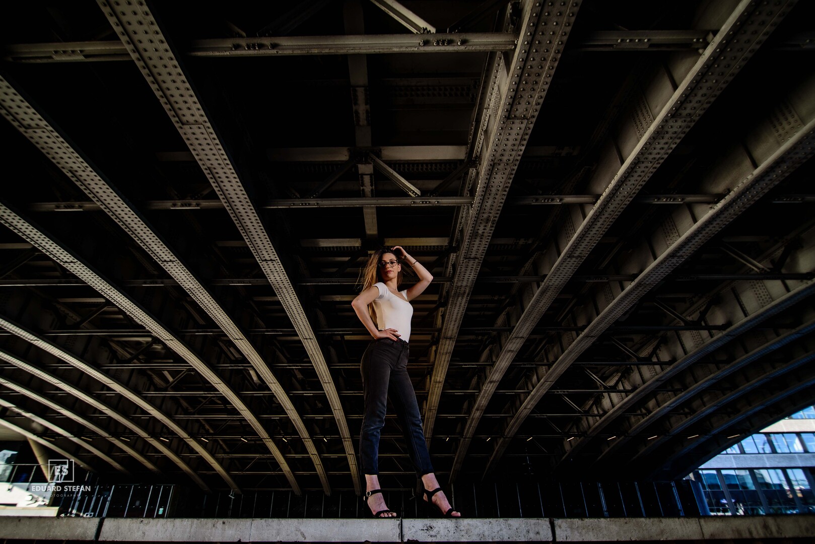 A stylish woman poses confidently beneath the steel beams of a modern bridge, embodying urban elegance.