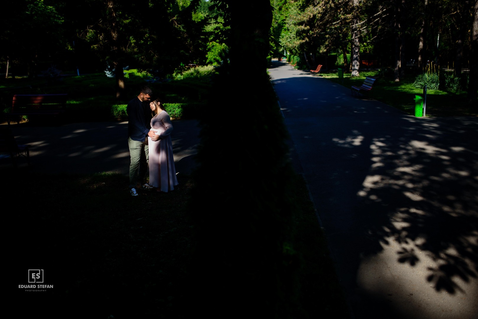 xpecting couple embracing in a quiet park, surrounded by trees and soft shadows from the afternoon light.