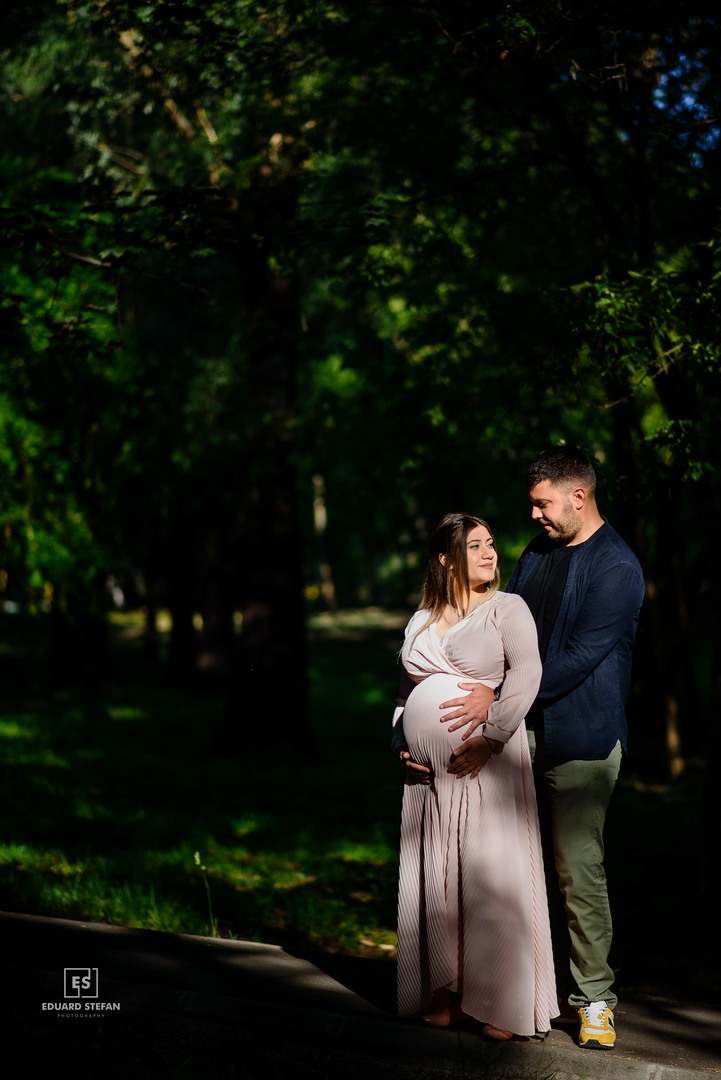 Pregnant woman in a flowing dress embraced by her partner in a sunlit forest, surrounded by lush greenery.