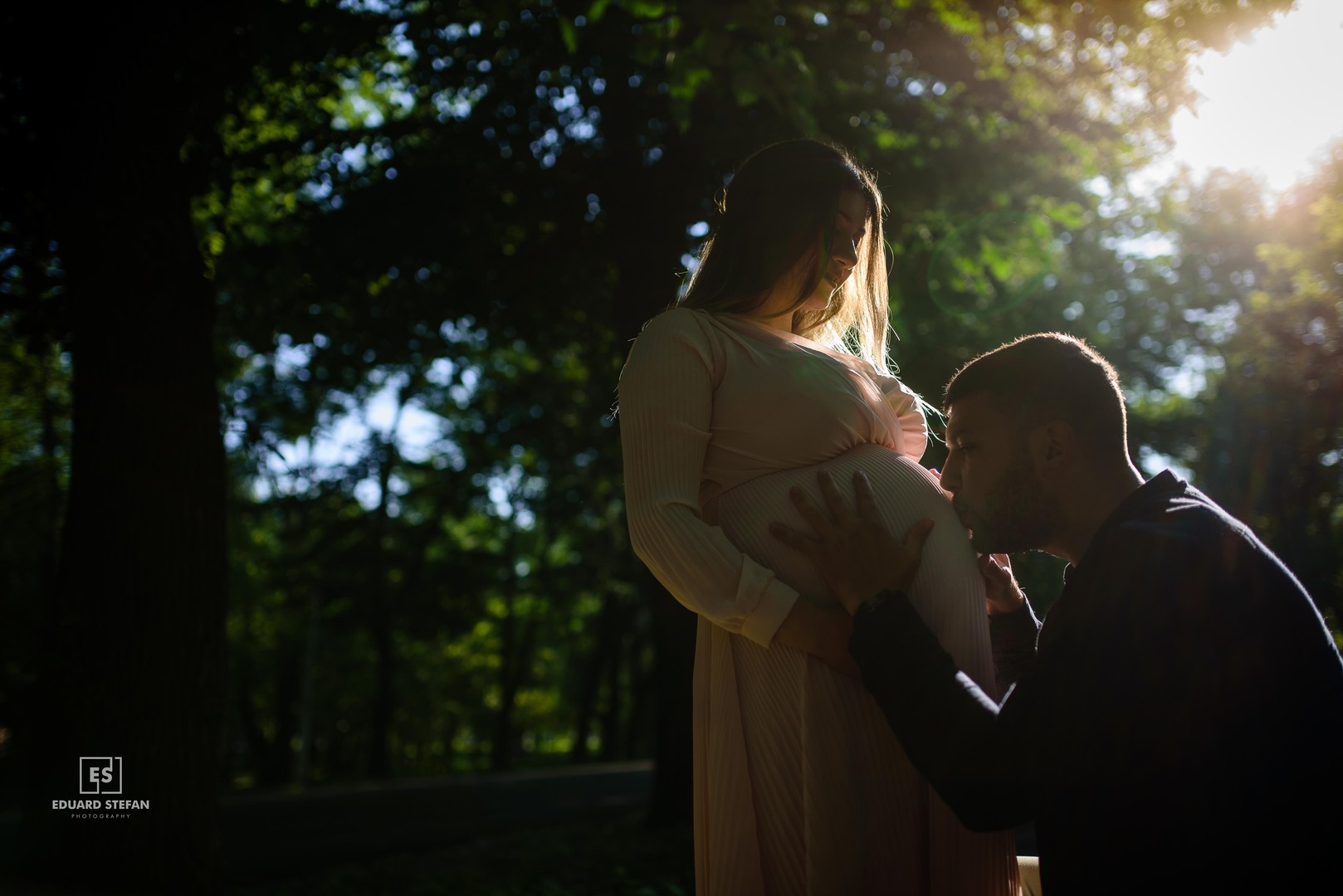 Man kissing the baby bump of a pregnant woman in a serene forest setting, illuminated by soft sunlight.