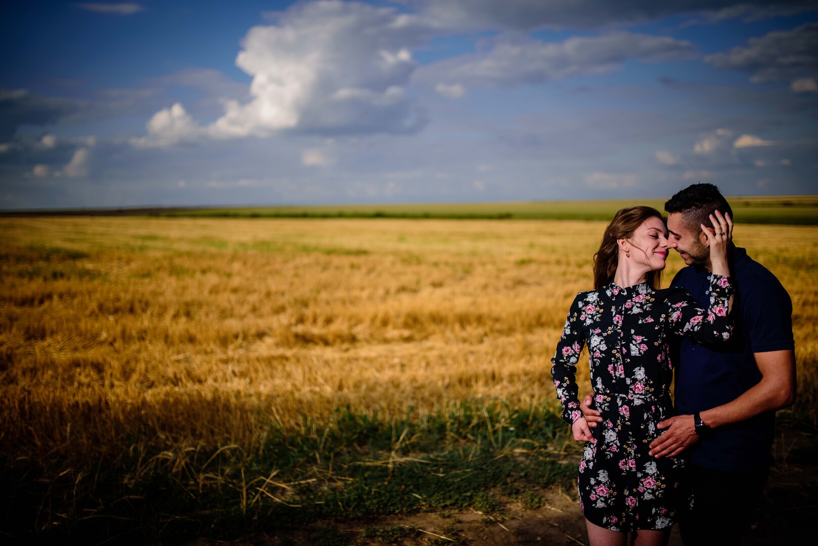 ouple embracing lovingly in a golden wheat field under a blue sky with scattered clouds.