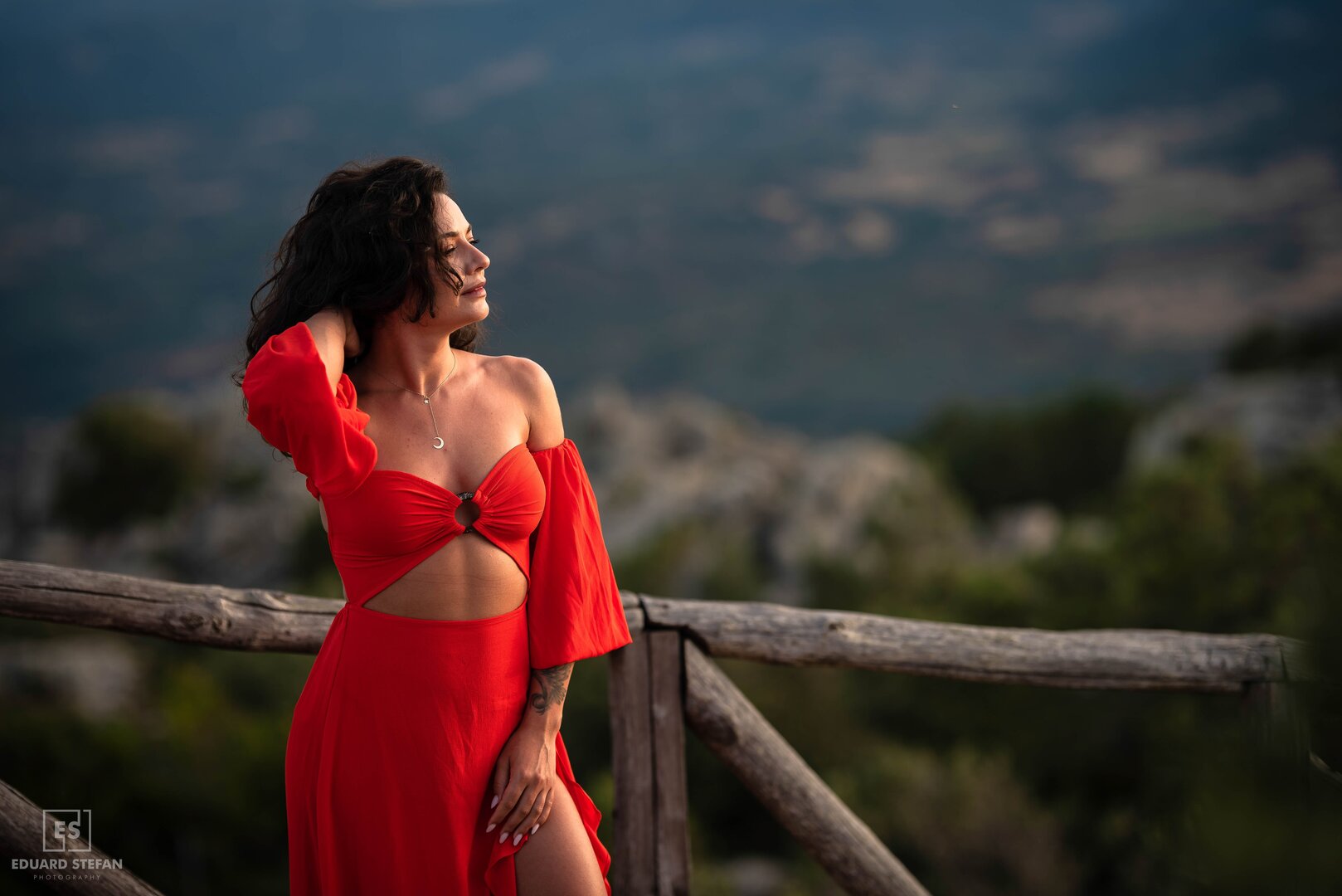 A woman in a bold red dress poses gracefully against a rustic wooden fence, with a serene mountainous landscape in the background.