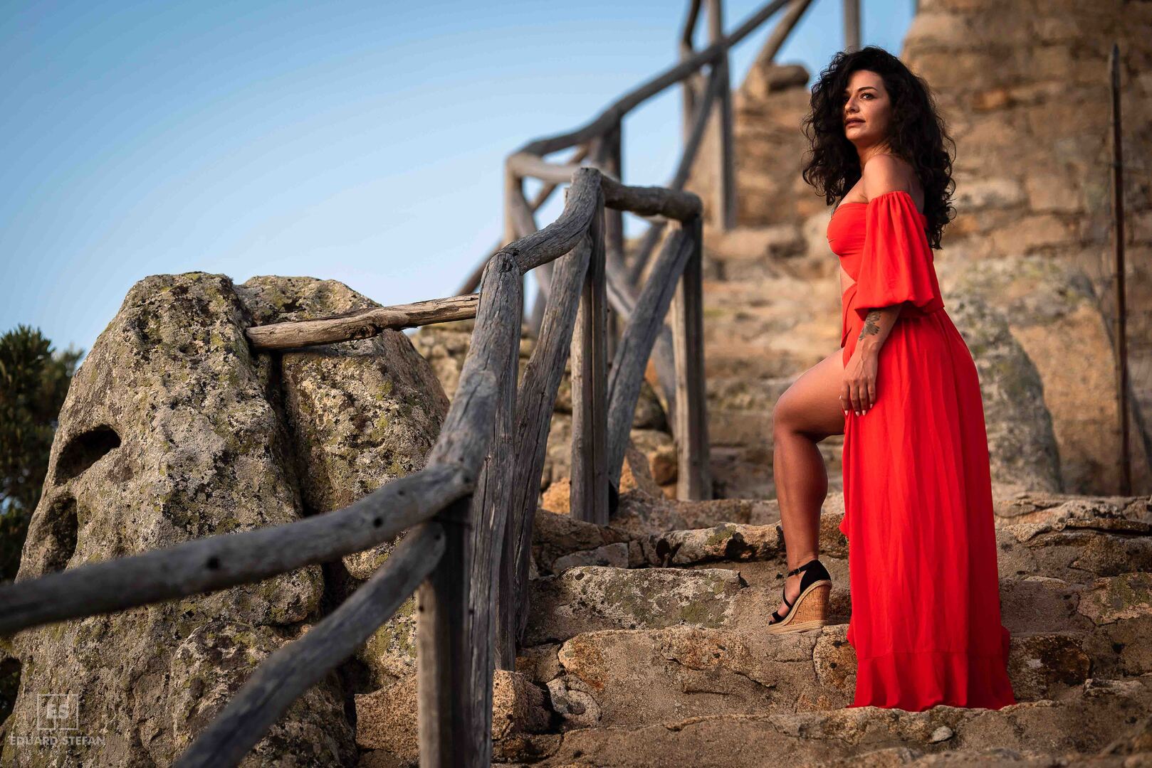 A confident woman in a flowing red dress stands on rustic stone steps, framed by a wooden railing and a clear blue sky, exuding elegance and strength.