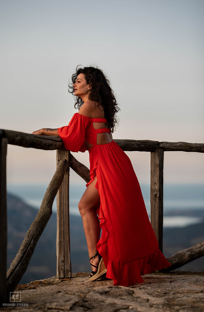 A striking woman in a vibrant red dress leans gracefully on a wooden railing, gazing into the distance with wind-tousled curls, set against a serene mountainous backdrop.