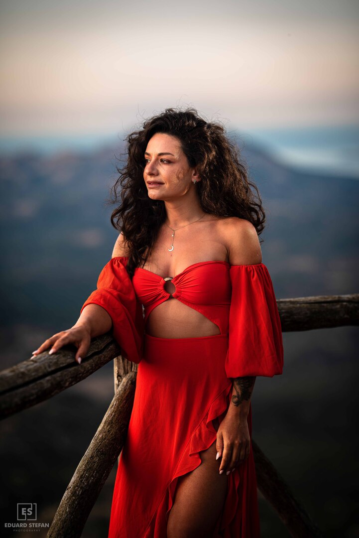 A poised woman dressed in a flowing red gown stands against a wooden railing, her serene expression reflecting the tranquil mountain view in the background.