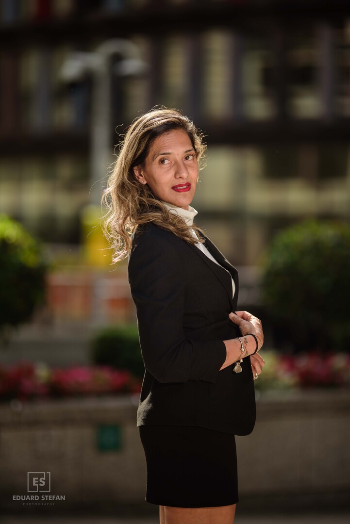 A poised woman glances confidently over her shoulder, dressed in a tailored black blazer and skirt, illuminated by soft urban lighting with a vibrant city backdrop.