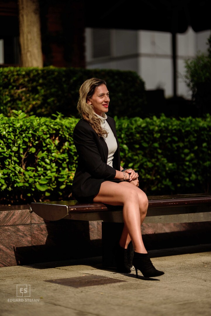 A woman sits gracefully on a polished bench, dressed in a black blazer and skirt, exuding poise against a lush green hedge background under warm sunlight.