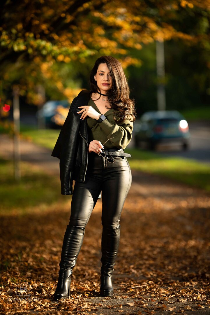 A confident woman stands amidst fallen autumn leaves on a sunlit pathway, dressed in sleek leather trousers and holding a black jacket over her shoulder, with golden foliage framing the scene.