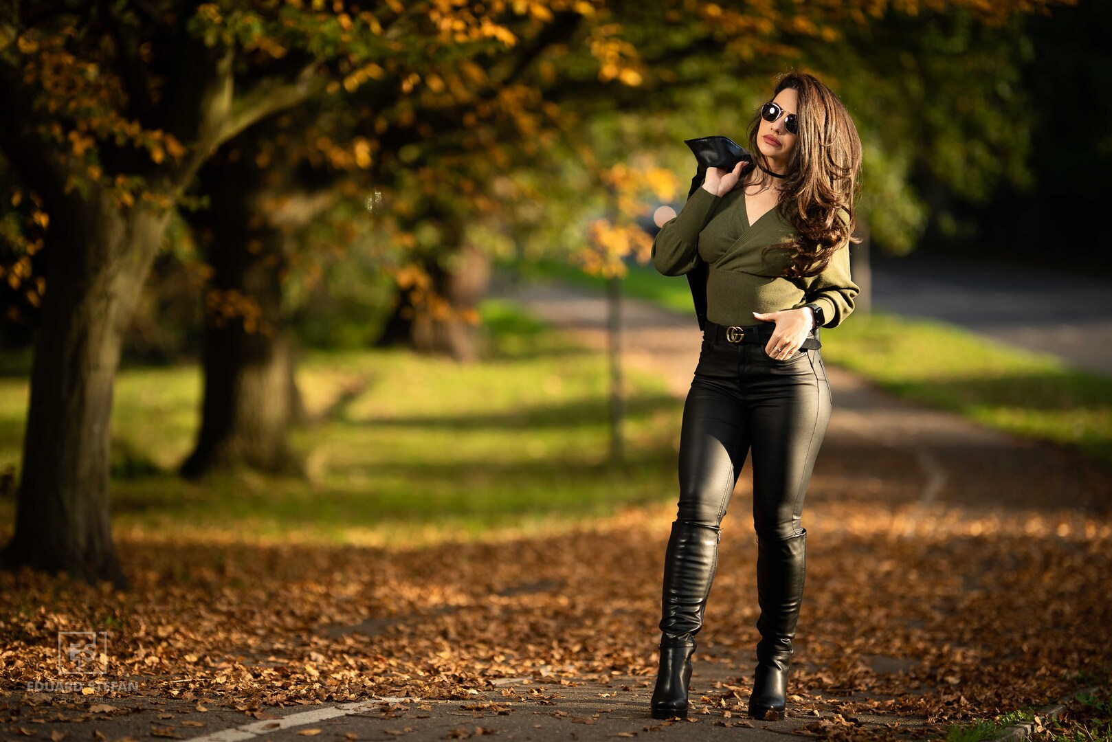 A stylish woman in leather trousers and sunglasses strolls through a sunlit, leafy path.