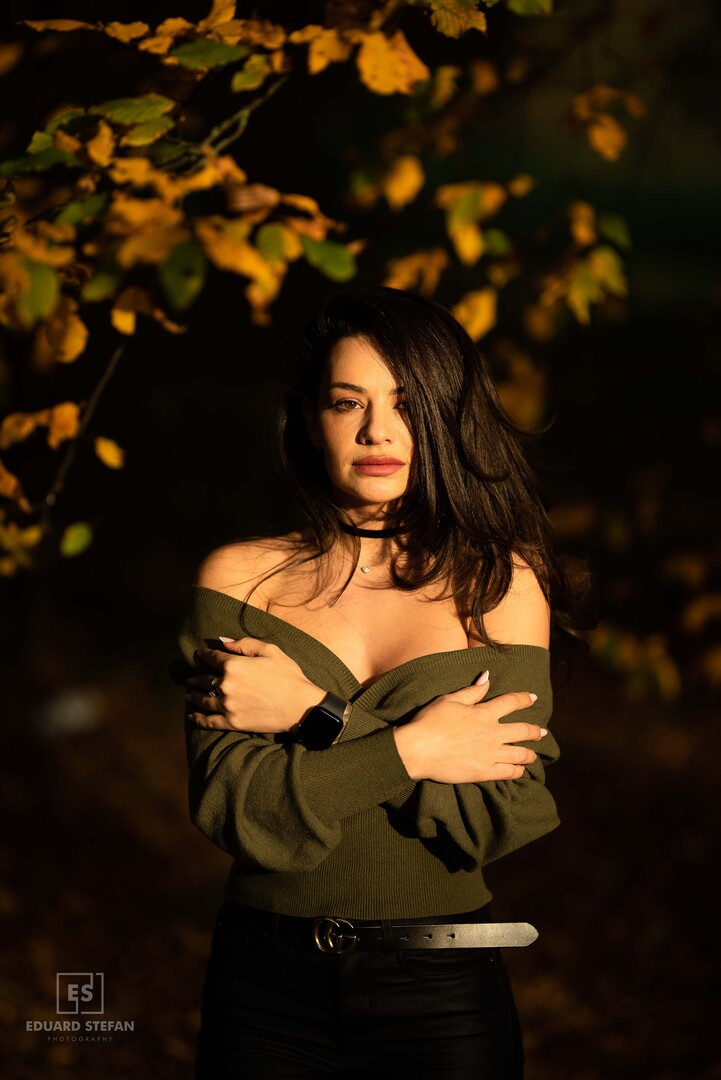 A woman in an off-shoulder sweater stands confidently under golden autumn leaves, illuminated by warm sunlight.