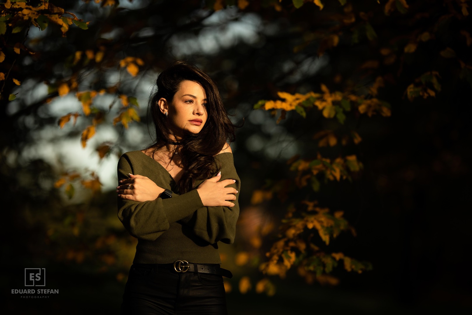 A woman stands gracefully under the golden glow of autumn foliage, her confident pose accentuating the soft light and vibrant leaves.