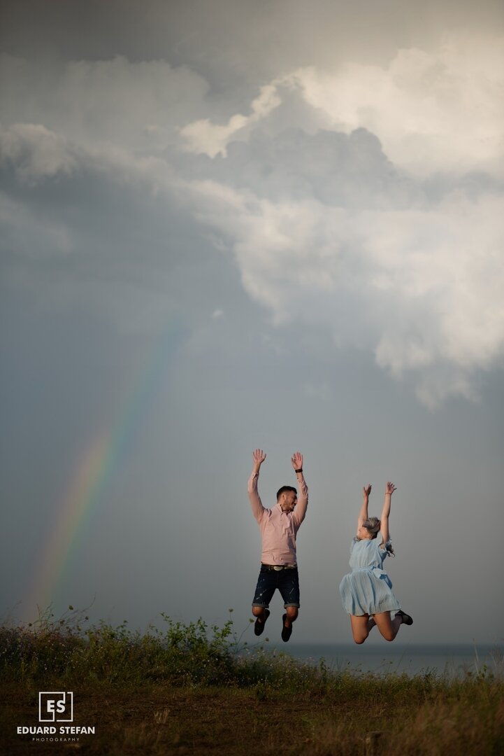 Joyful couple jumping in the air on a hilltop, with a rainbow and dramatic cloudy sky in the background.