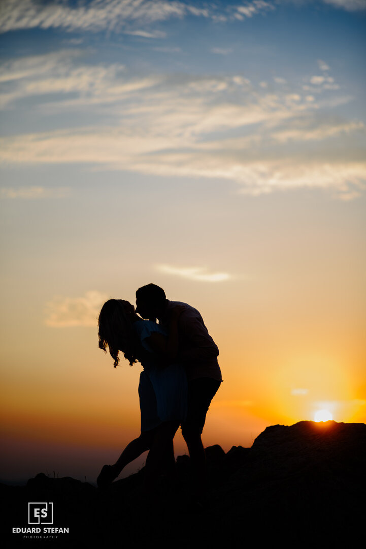 Silhouetted couple sharing a romantic kiss on a hilltop during a vibrant sunset with a colourful sky.