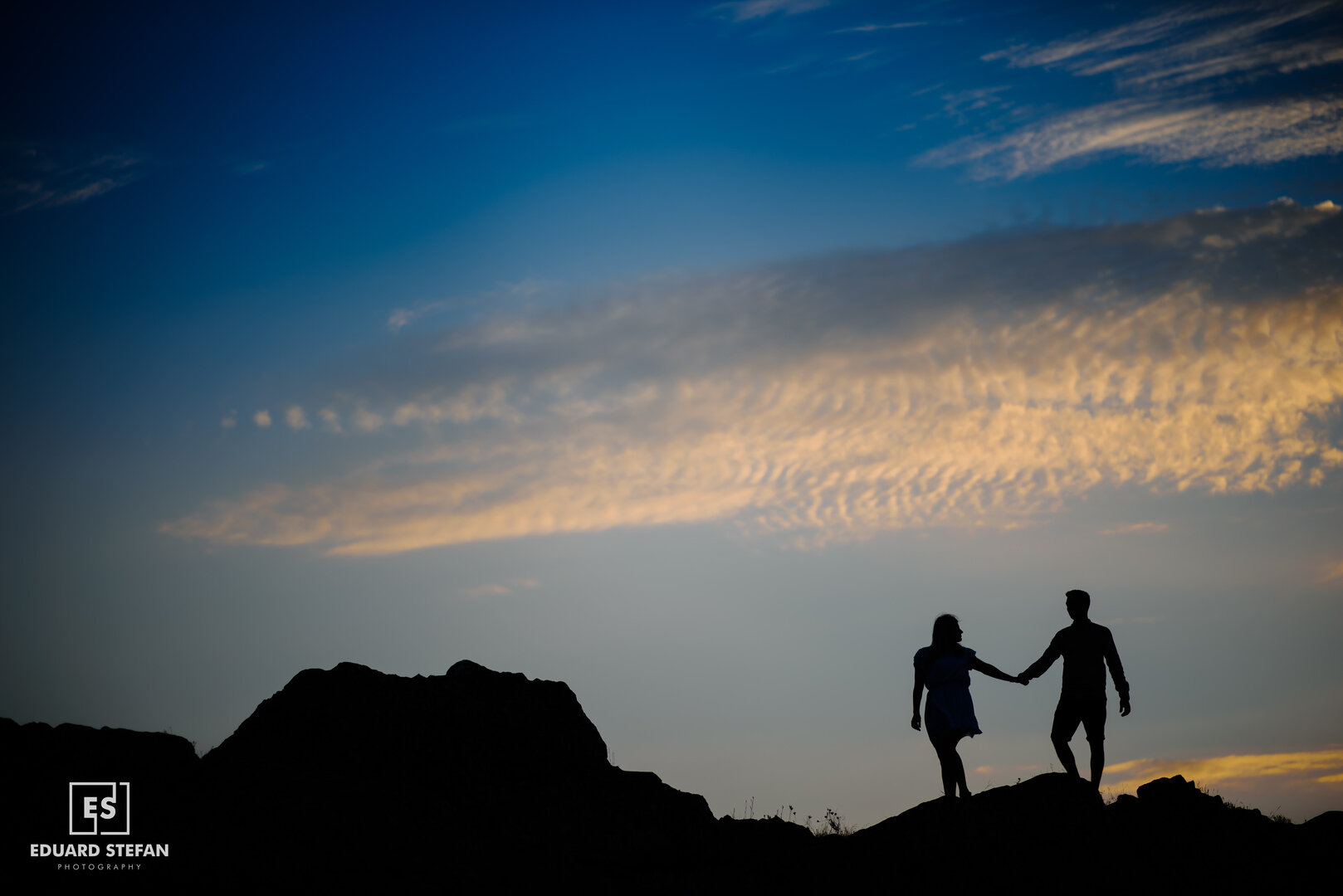 Silhouetted couple holding hands while walking on a hilltop during a vibrant sunset with a dramatic sky.