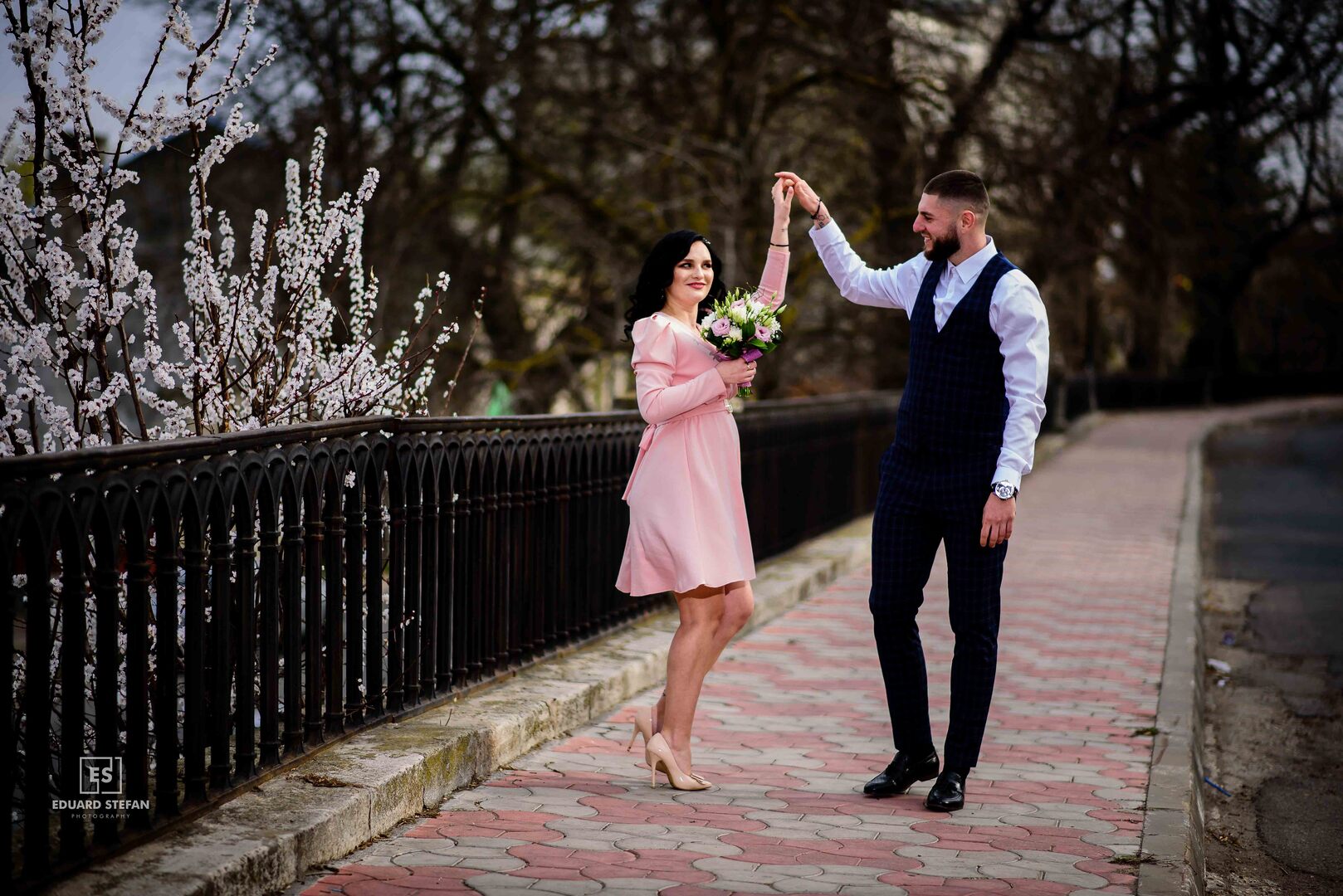 Man in a formal suit twirling a woman in a pink dress holding a bouquet, walking along a blooming spring pathway.