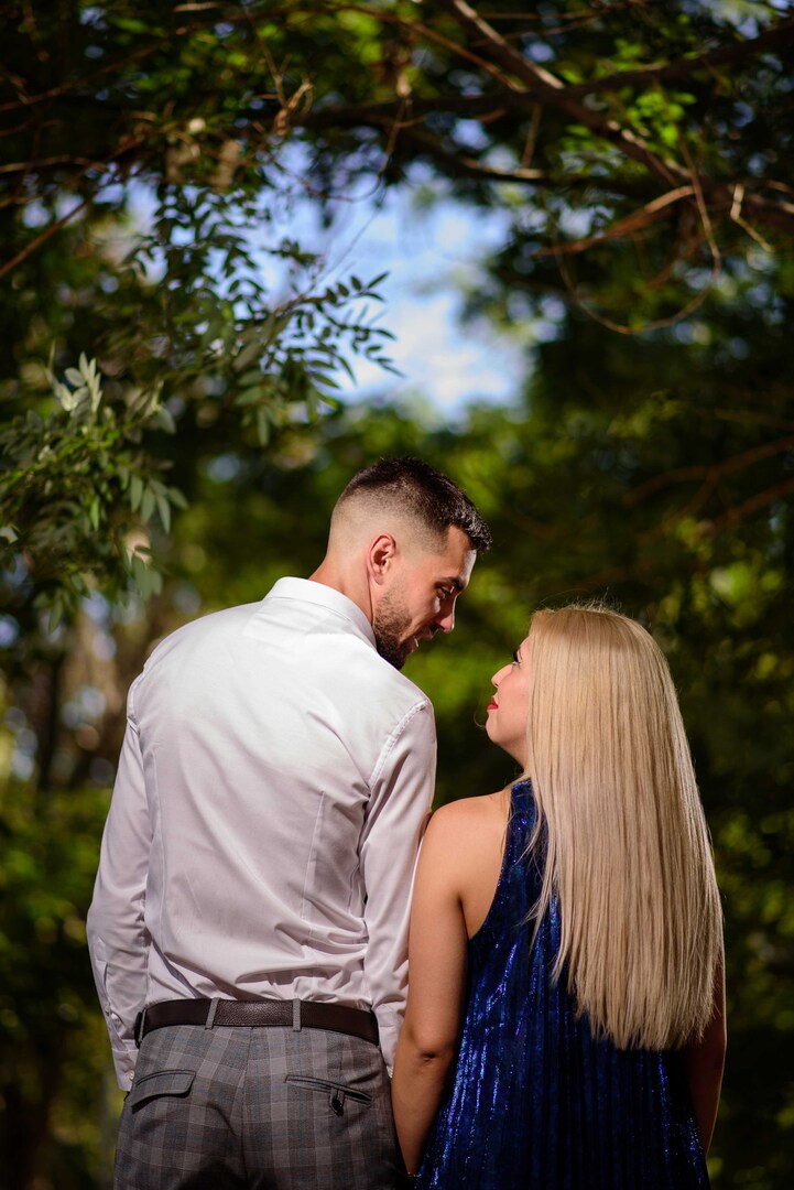 Couple standing close together in a forest, gazing at each other under a canopy of green leaves.