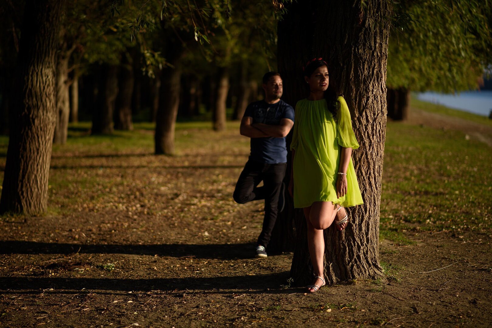 Woman in a vibrant green dress leaning against a tree, with a man standing in the background under the warm glow of sunlight.