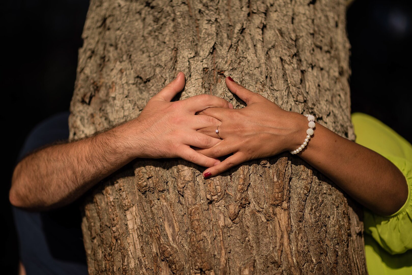 lose-up of a couple's hands holding each other around a tree trunk, showcasing a ring and bracelet.