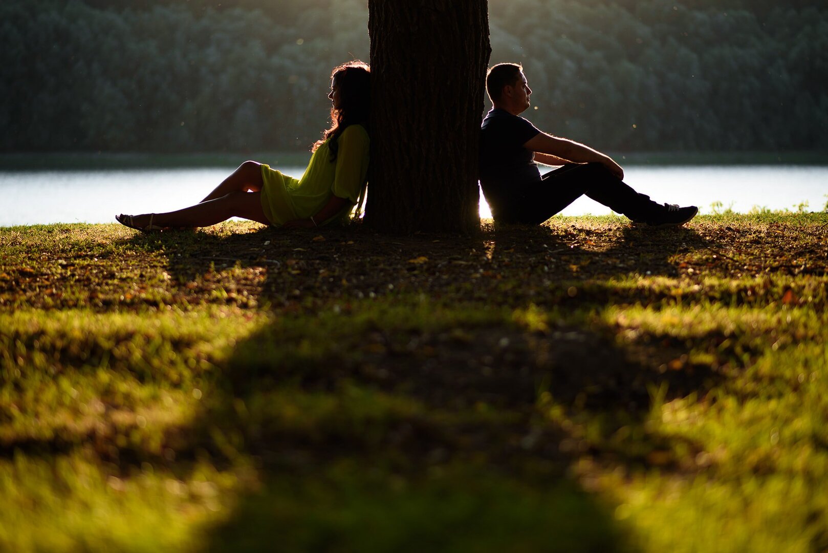 Couple sitting back-to-back against a tree by a serene lake, bathed in warm evening sunlight.