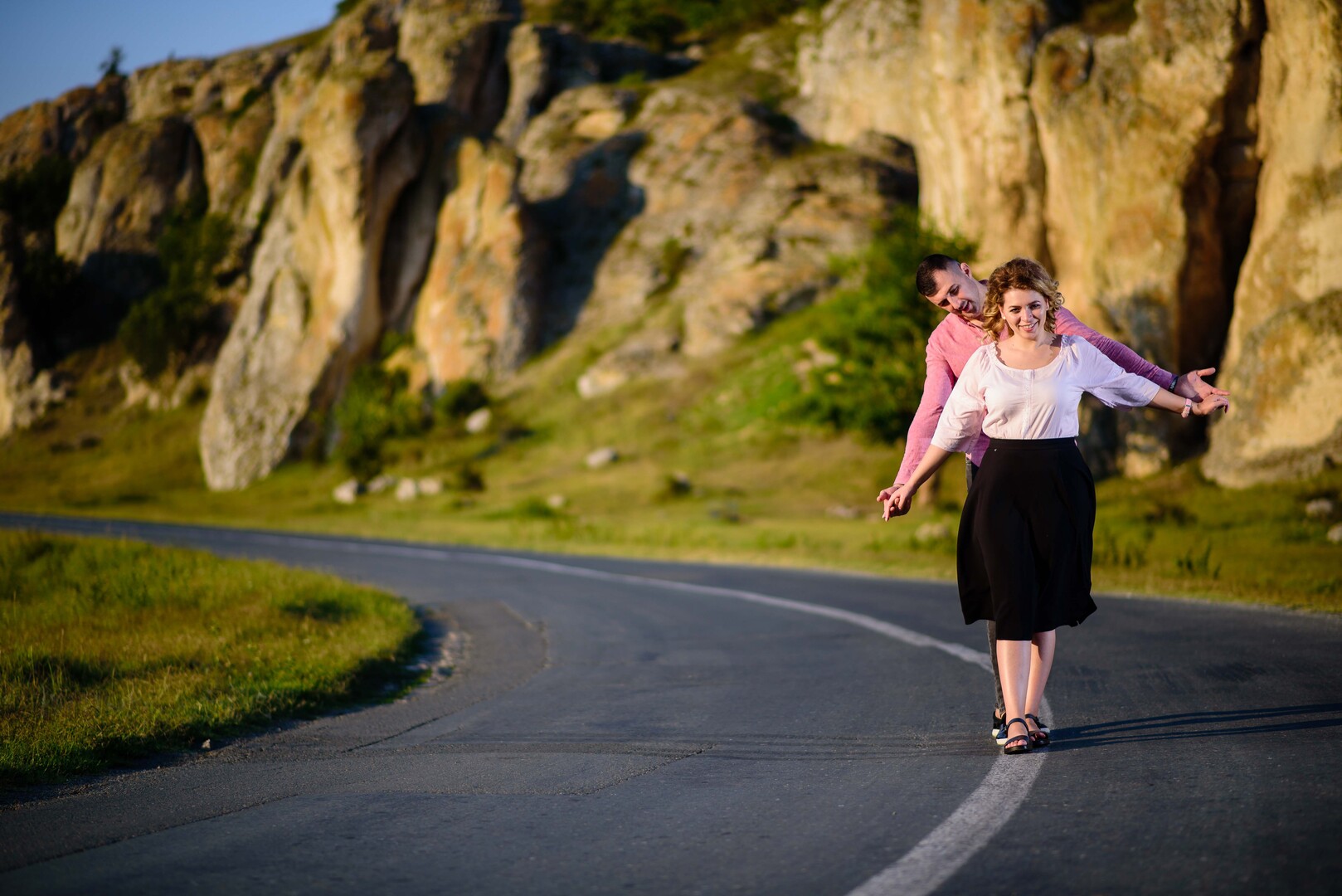 Couple playfully balancing on a winding road with rocky cliffs in the background, enjoying a sunny day outdoors