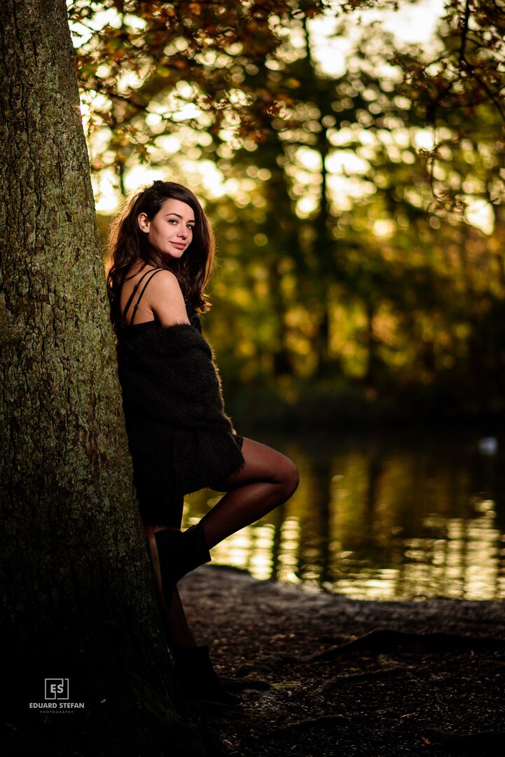 A woman leans casually against a tree by a serene lakeside, her confident expression complementing the golden hues of the setting sun filtering through the forest.