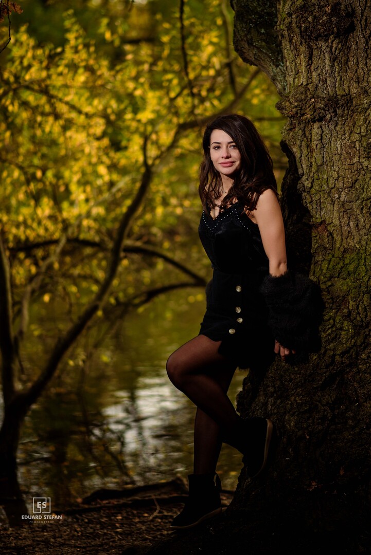 A woman stands gracefully against a tree, her dark attire contrasting beautifully with the vibrant autumn leaves reflecting on the tranquil lake in the background.